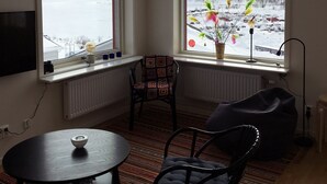 A room with a large window offering a view of a snowy landscape and mountains. There is a chair, a table with a bowl, and a lamp on the windowsill. - RIBO Apartment Riksgränsen (Riksgränsen)