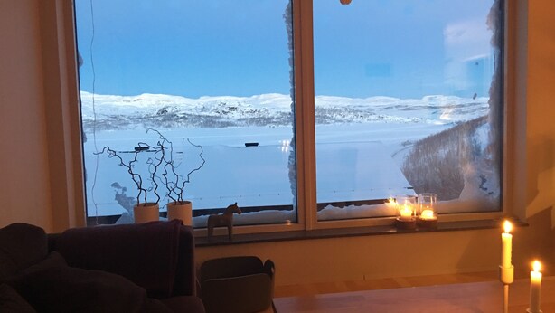 A room with a large window overlooking a snowy landscape. There are two potted plants on the windowsill, a candle holder with candles, and a dark-colored sofa. - RIBO Apartment Riksgränsen (Riksgränsen)
