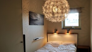 A neatly made bed with a headboard, a wall-mounted picture, a unique ceiling light fixture, and a window with a view of a building. - RIBO Apartment Riksgränsen (Riksgränsen)