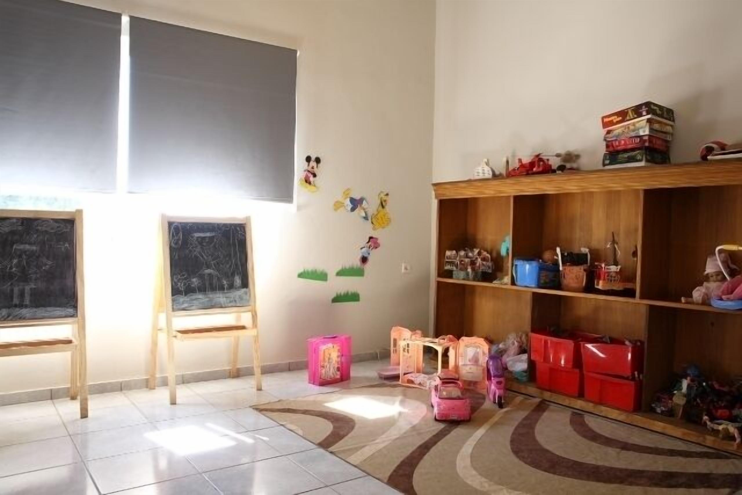 A classroom with two chalkboards, a wooden shelf with books and toys, and storage bins on the floor.