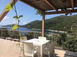 A terrace with a white table and chairs, overlooking a coastal landscape with hills and a body of water.