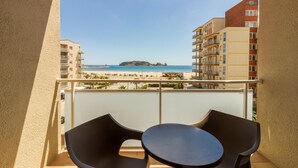Ein Balkon mit zwei Stühlen und einem Tisch, der Blick auf einen Strand mit Gebäuden und einer kleinen Insel im Hintergrund bietet.