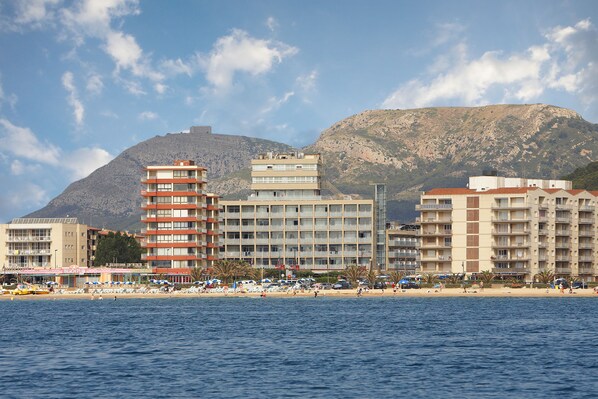 Un hotel frente a la playa con varios pisos, una playa en primer plano y una montaña al fondo.