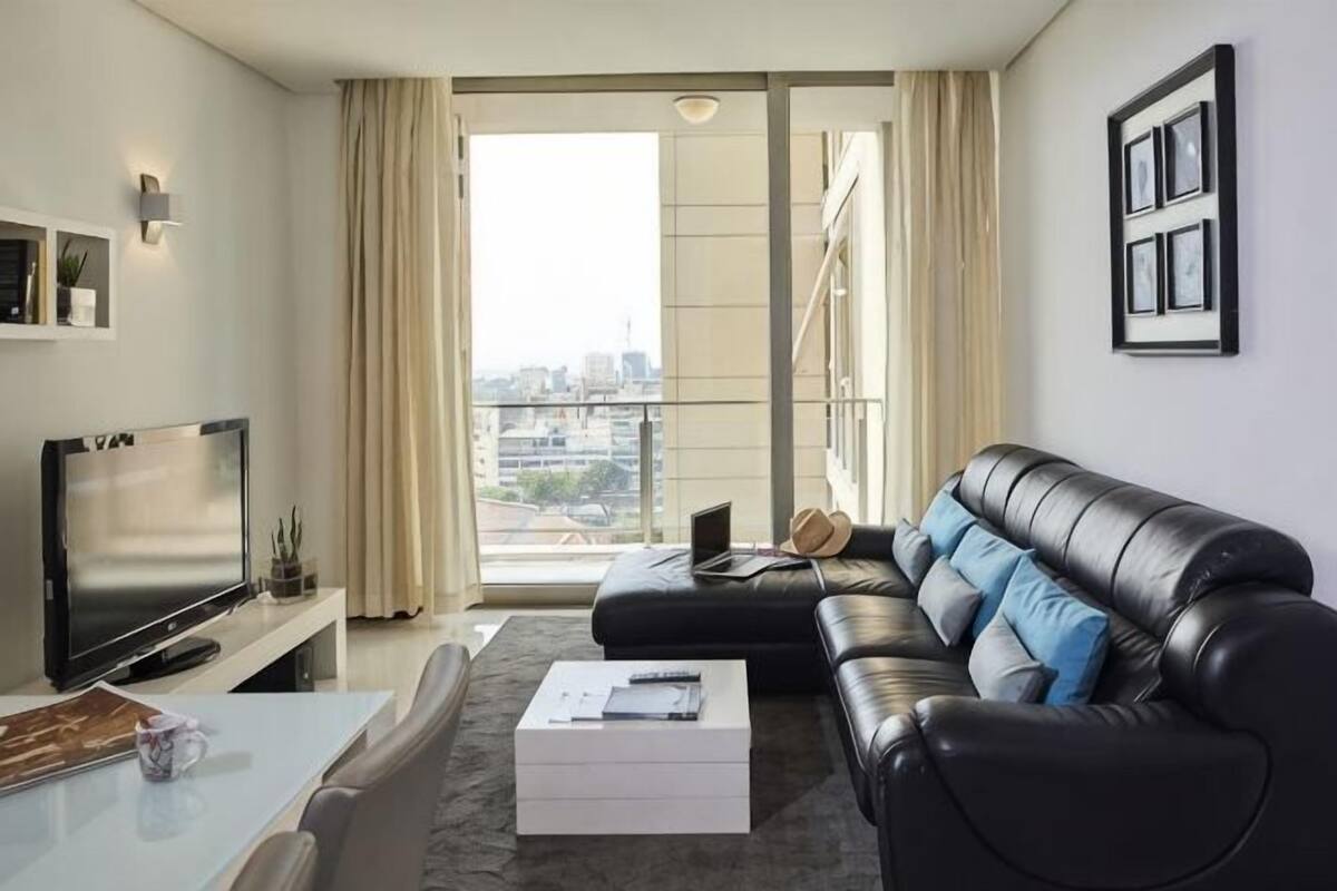 Una sala de estar moderna con un sofá de cuero negro, un televisor de pantalla plana, una mesa de comedor y un amplio ventanal con cortinas.