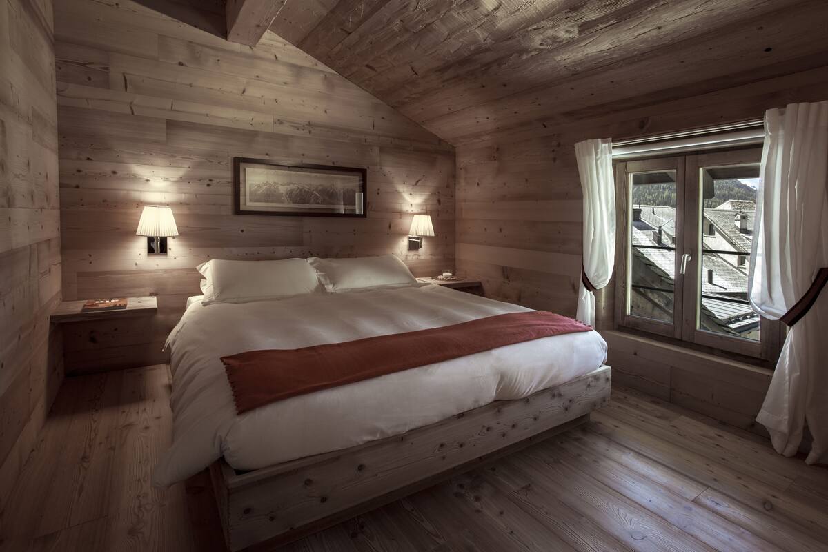 A wooden cabin bedroom with a bed, two bedside lamps, a window with curtains, and a framed picture on the wall.