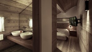 A wooden cabin interior with a wooden ceiling, a large mirror, two sinks, a bed, and a mounted TV. - Chesa Stuva Colani (Madulain)