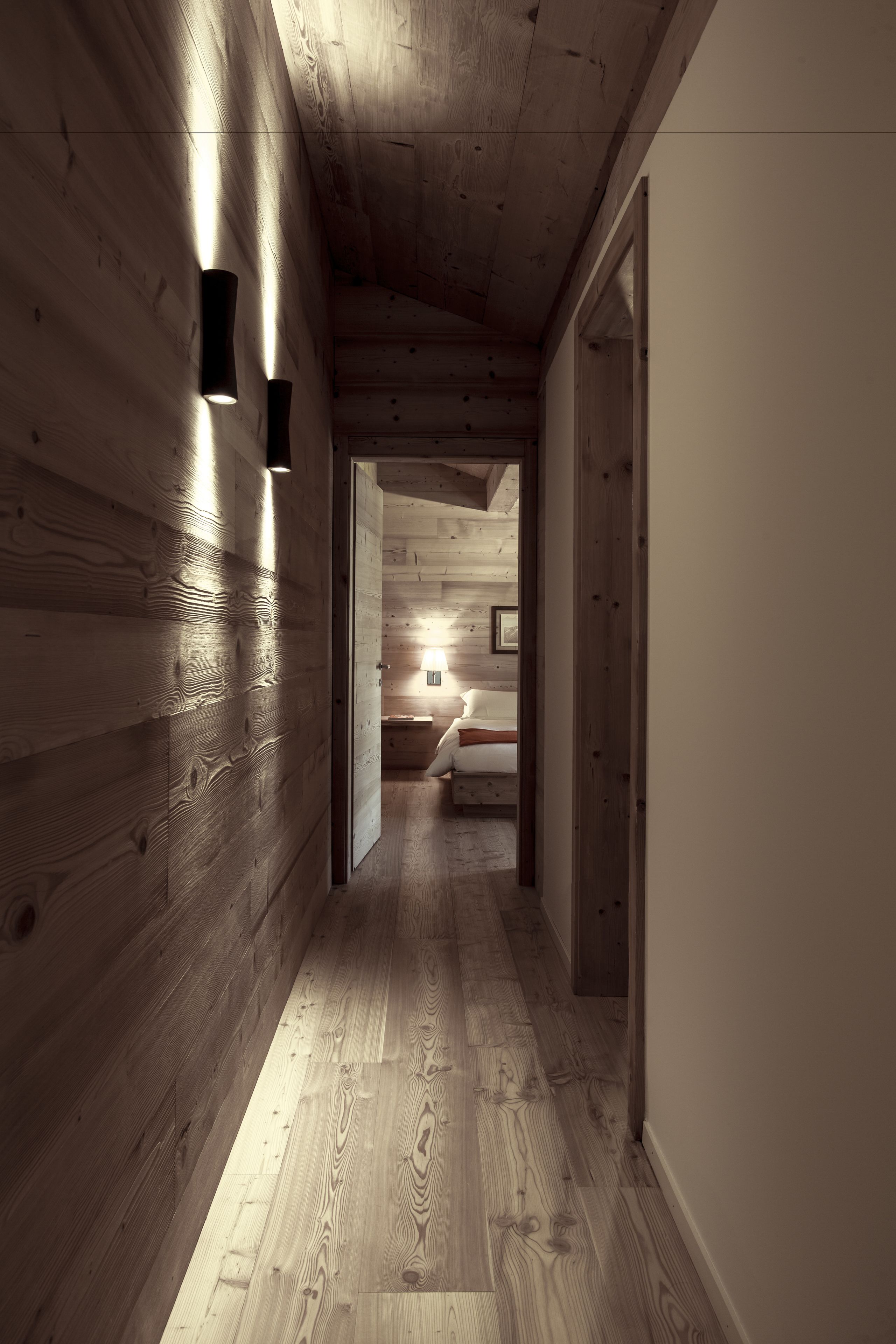 A wooden hallway with a bed in the distance, a nightstand, and a lamp.