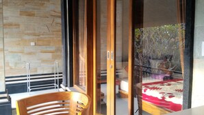 A room with a wooden chair and table, a stone wall, and a sliding glass door leading to a balcony with a table and chairs. - Rumah Ary Homestay & Spa (Ubud)