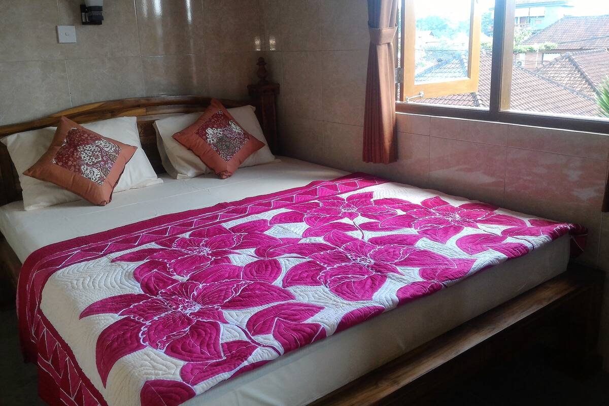 A bed with a floral bedspread, two pillows, a bedside table, and a window with curtains.