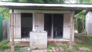 A rustic outdoor structure with a metal roof and open sides, featuring a concrete water dispenser and a toilet. - KK Hut (Pai)