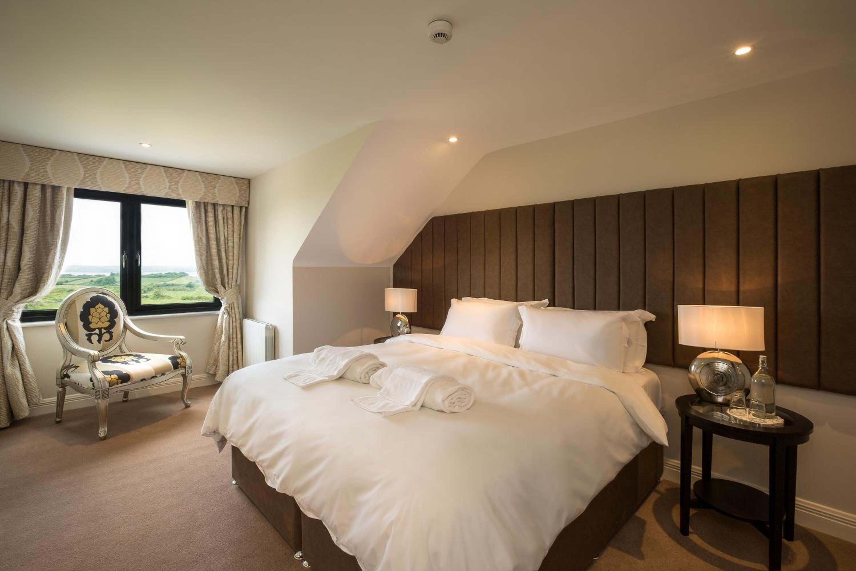 Una habitación de hotel con una cama grande, dos mesitas de noche, una silla y vistas al exterior a través de una ventana.