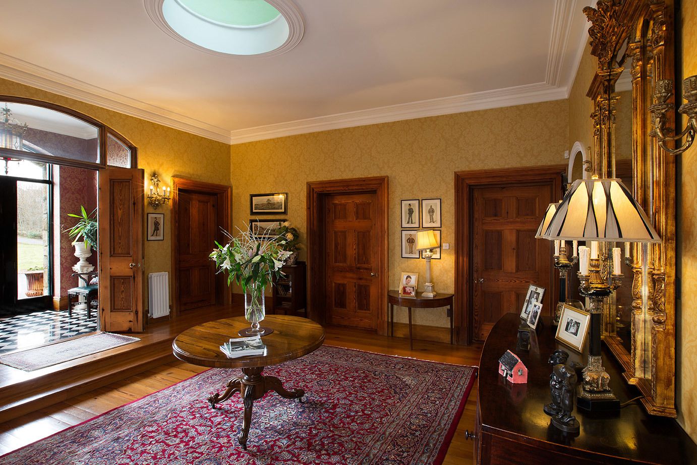 A grand room with a large wooden door, a round table with a vase of flowers, a patterned rug, and a chandelier.
