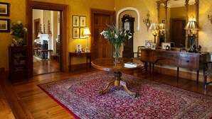 Una habitación formal con piso de madera, un tapete rojo estampado, una mesa redonda con un jarrón de flores, un escritorio de madera con cajones y un espejo grande.