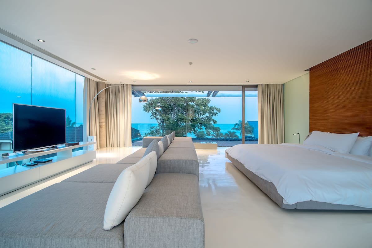 A modern bedroom with a large bed, a sofa, a TV, and a balcony with a view of the ocean.