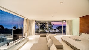A modern hotel room with a large TV, a sofa, a bed, and a balcony with a view of the sunset.