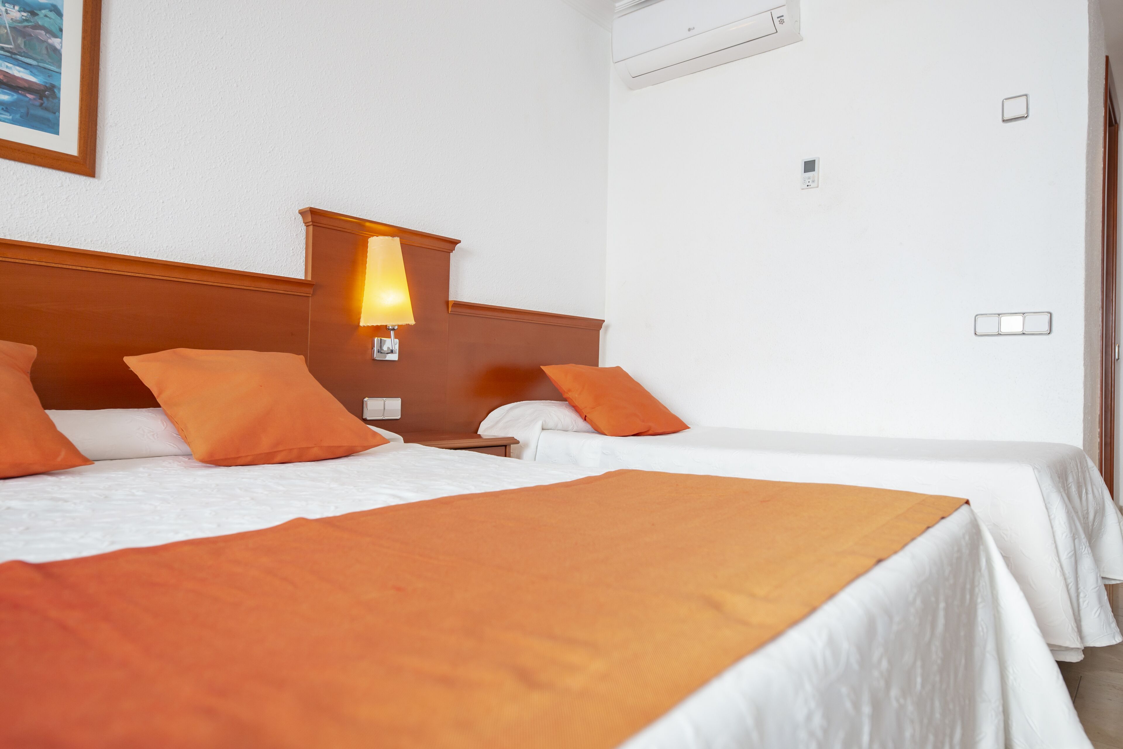 A hotel room with two beds, each with white bedding and orange accents, a nightstand with a lamp, and a painting on the wall.