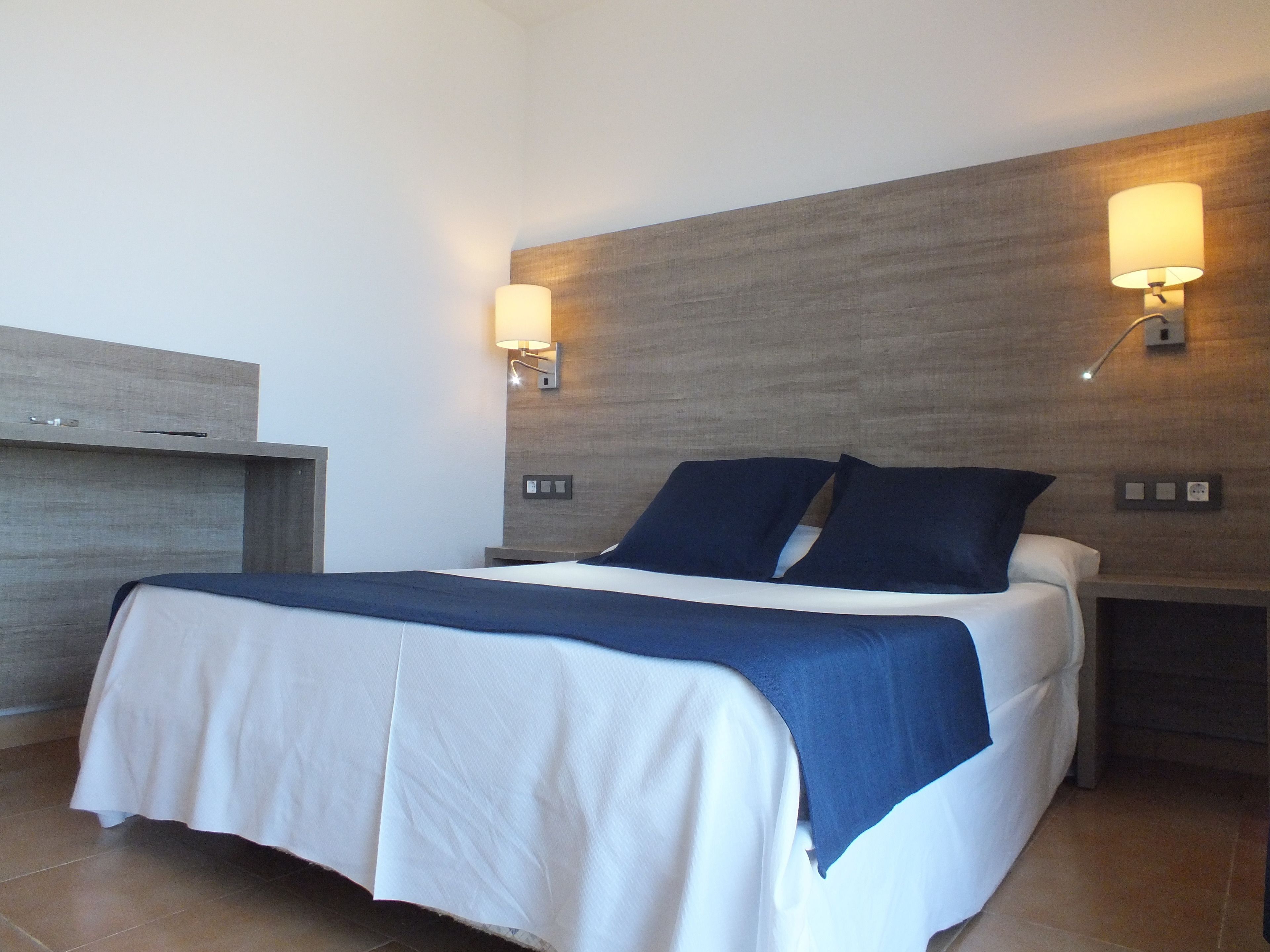 A neatly made bed with a white and blue bedspread, two wall-mounted lamps, and a wooden headboard.