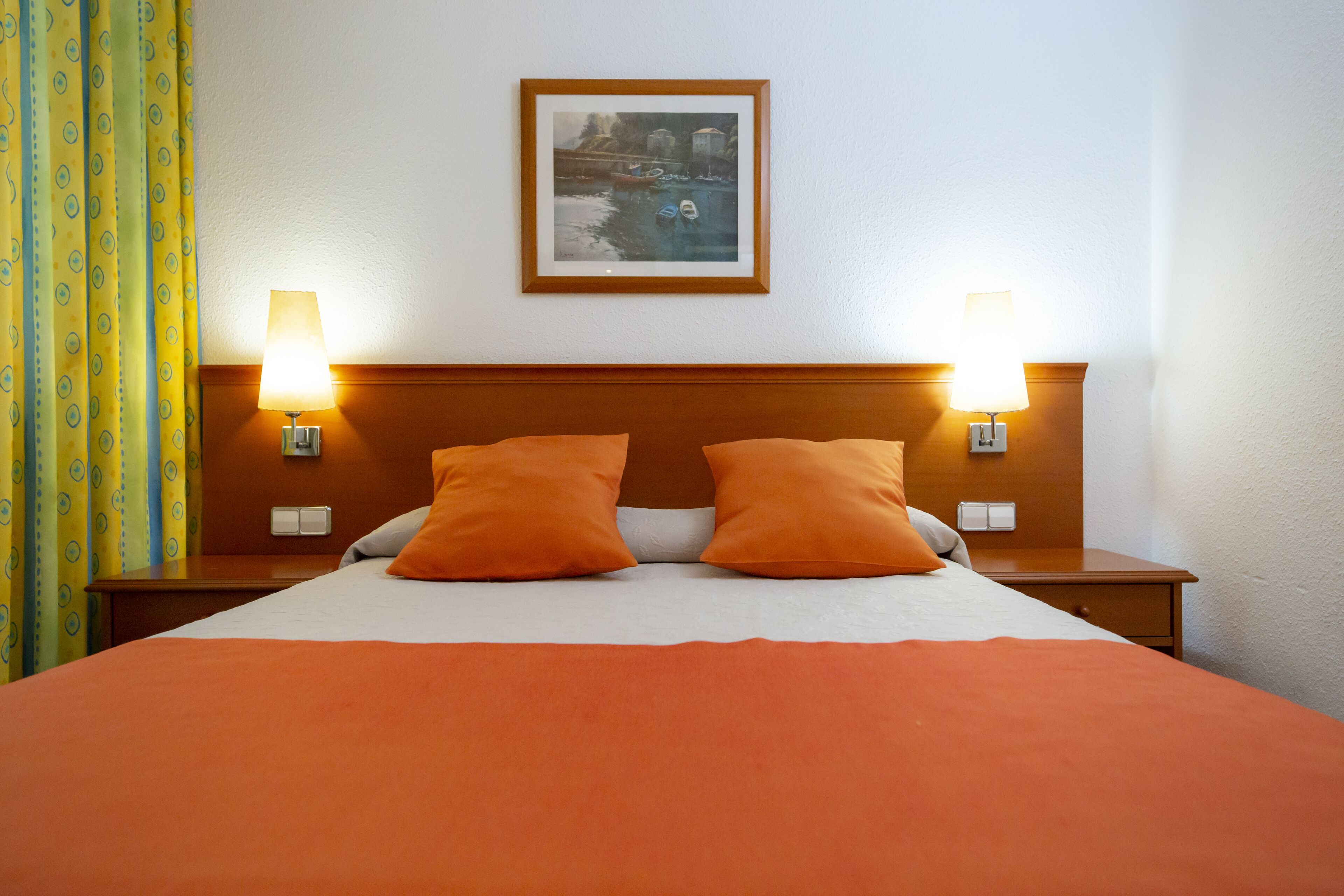A hotel room with a large bed, two orange pillows, a wooden headboard, two bedside lamps, and a framed picture on the wall.
