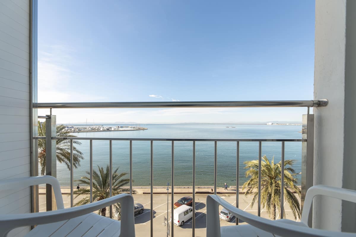 Un balcón con vista al mar, palmeras y coches aparcados.