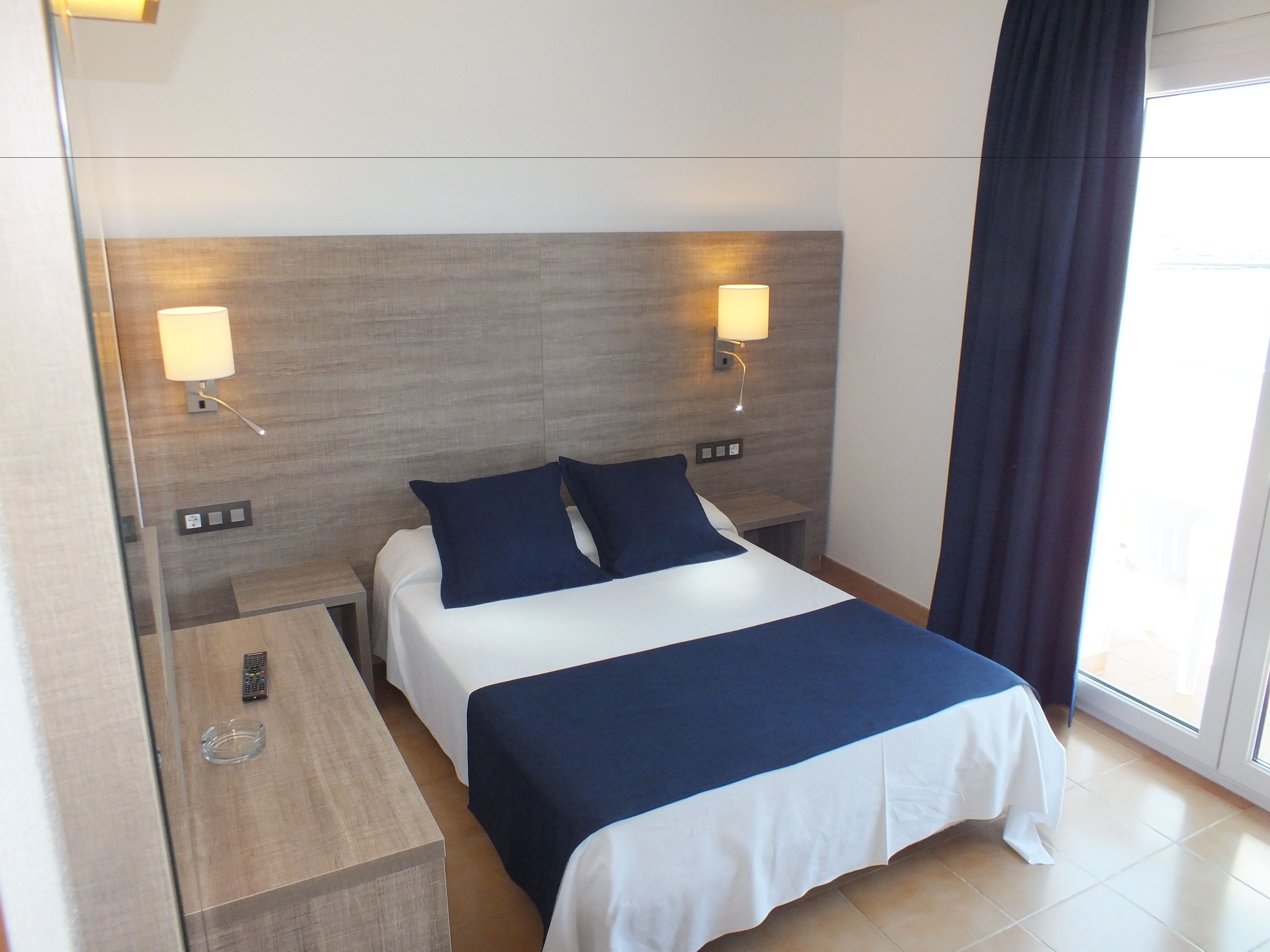 Une chambre d’hôtel avec un lit, deux tables de chevet, une lampe sur chaque table et une fenêtre avec un rideau.