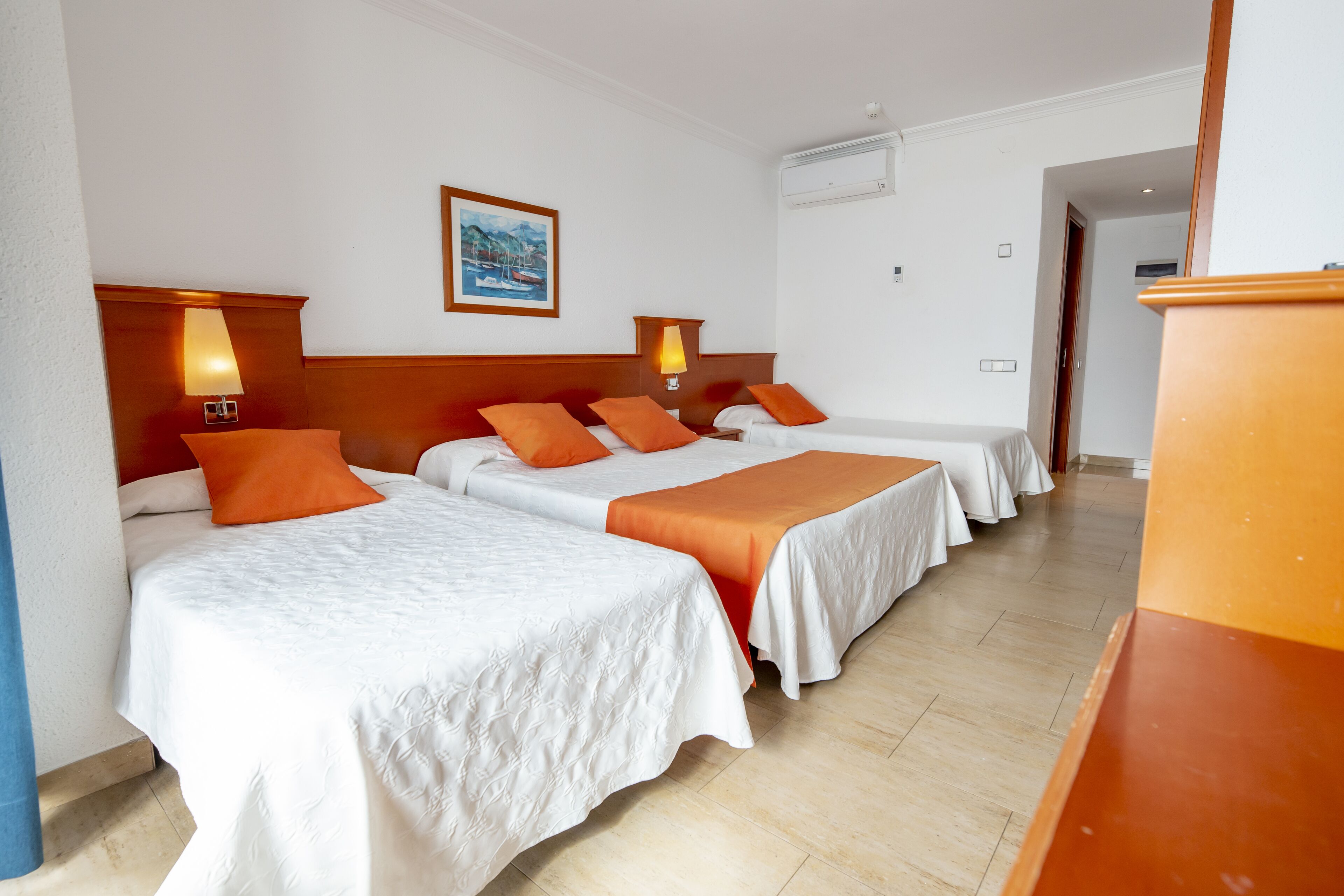 A hotel room with three beds, each with white bedding and orange throw pillows. There are wall-mounted lamps on either side of the beds, and a painting above the headboard.