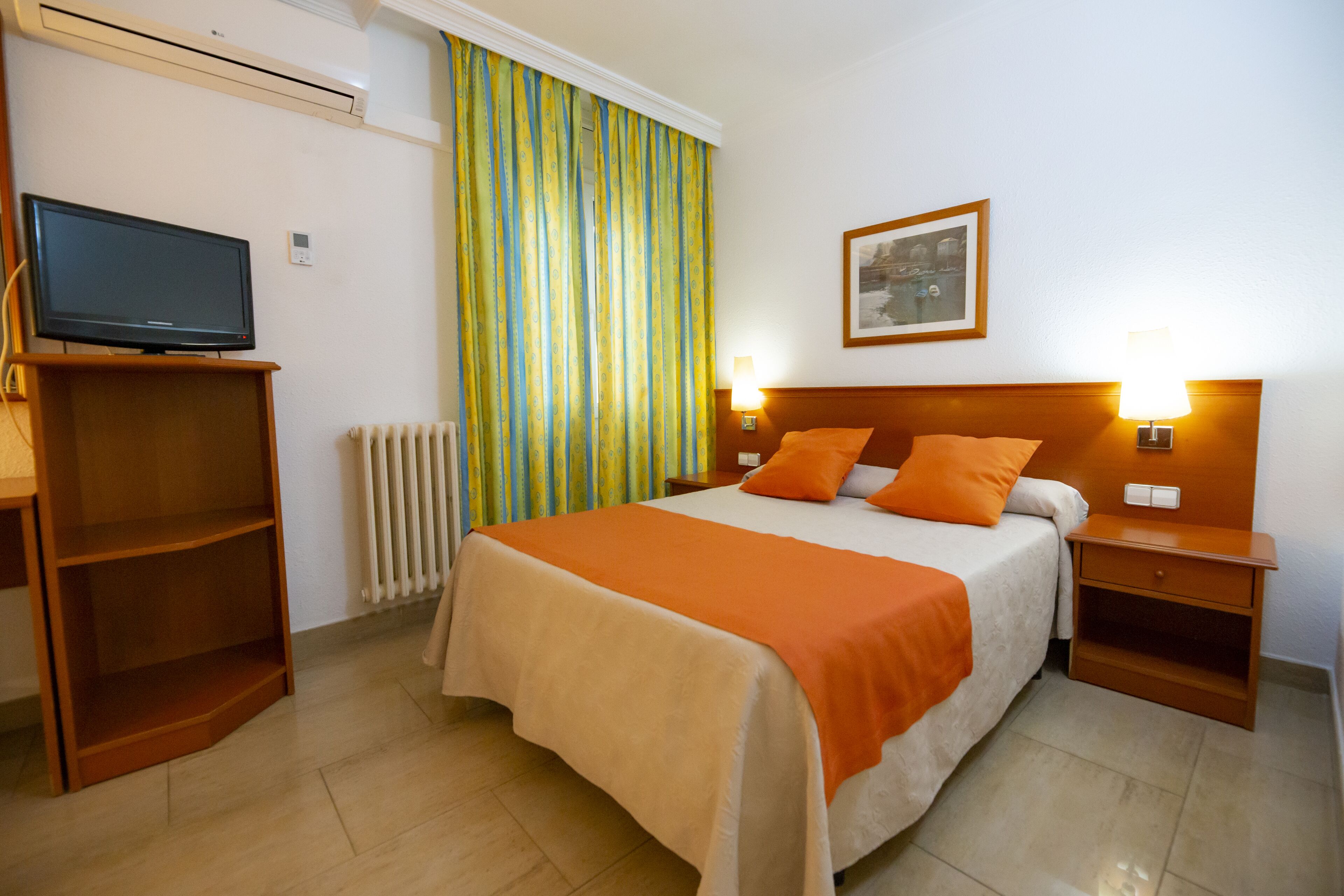 Habitación de hotel con cama, televisor sobre un mueble bajo, mesita de noche con lámpara y ventana con cortinas.