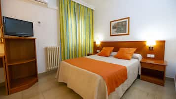 Habitación de hotel con cama, televisor sobre un mueble bajo, mesita de noche con lámpara y ventana con cortinas.