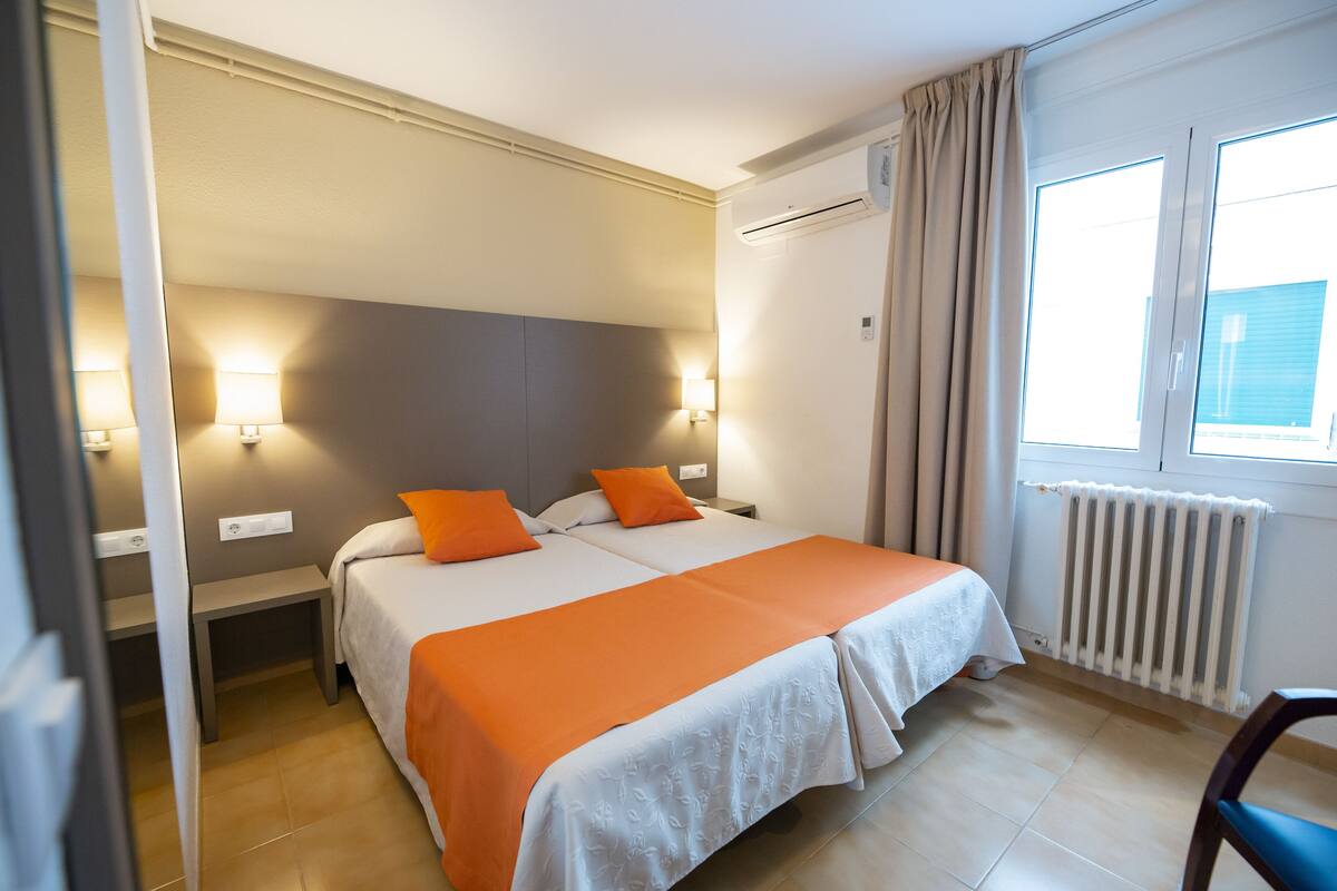 Una cama doble con ropa de cama naranja y blanca, una mesita de noche, una lámpara de pared y una ventana con cortinas.