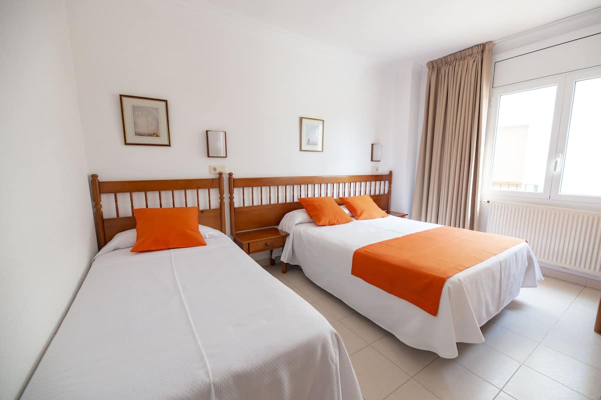 Habitación de hotel con dos camas individuales, cada una con ropa blanca y detalles naranja, un cabecero de madera, una mesita de noche y una ventana con cortinas.