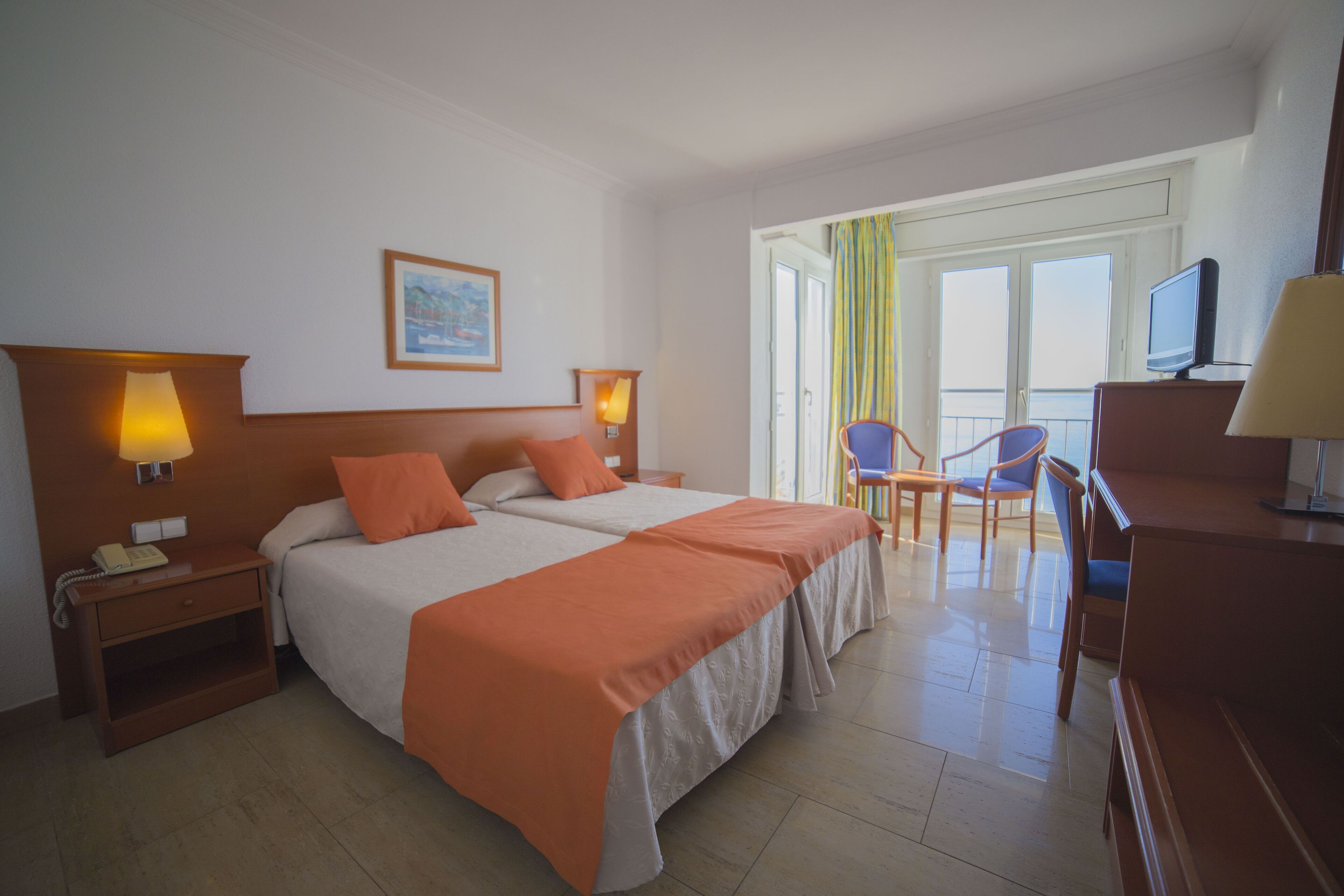 A hotel room with two beds, a desk, a chair, a TV, and a balcony with a view.