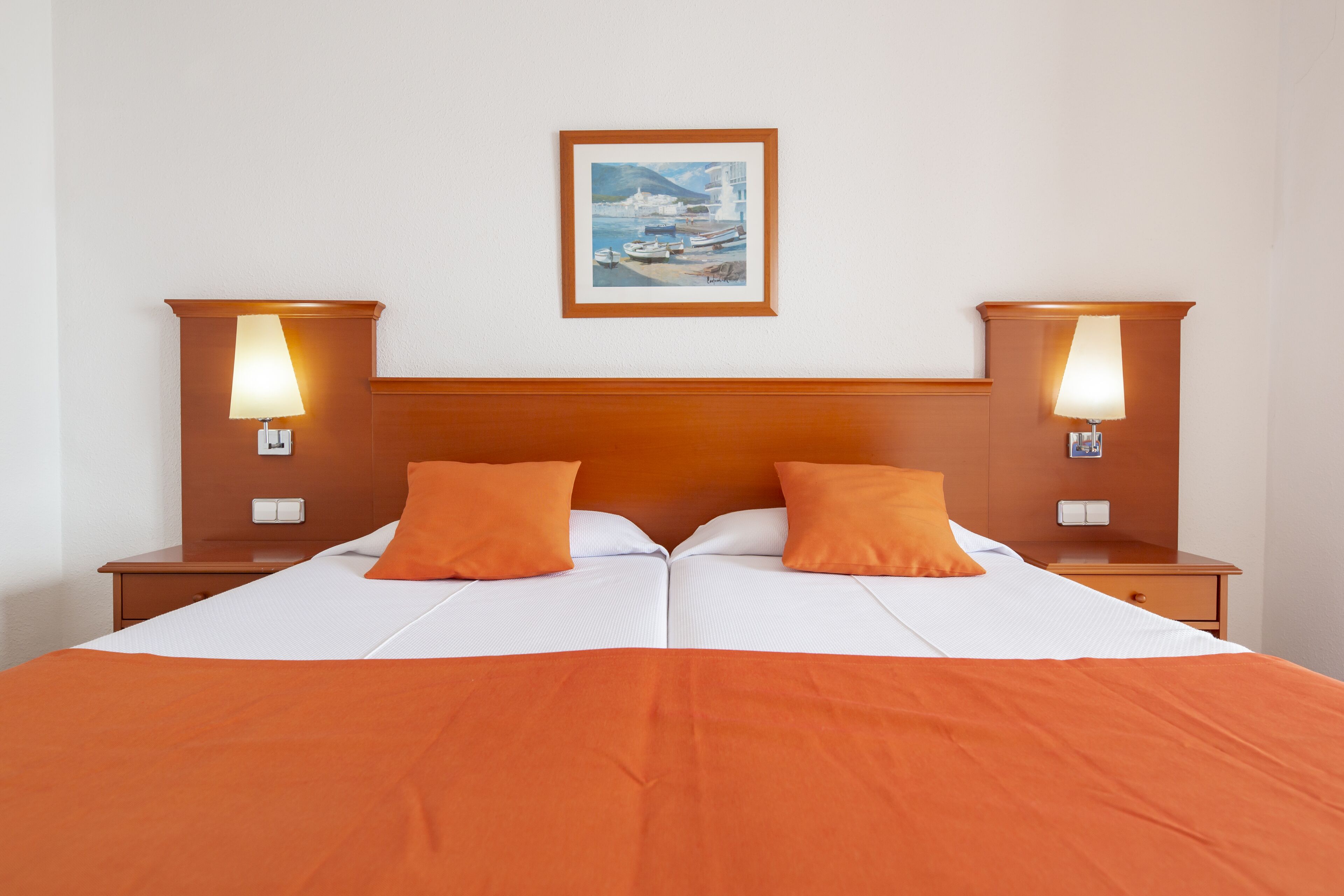 A double bed with an orange bedspread and two matching pillows, flanked by nightstands with lamps, and a framed painting above the bed.