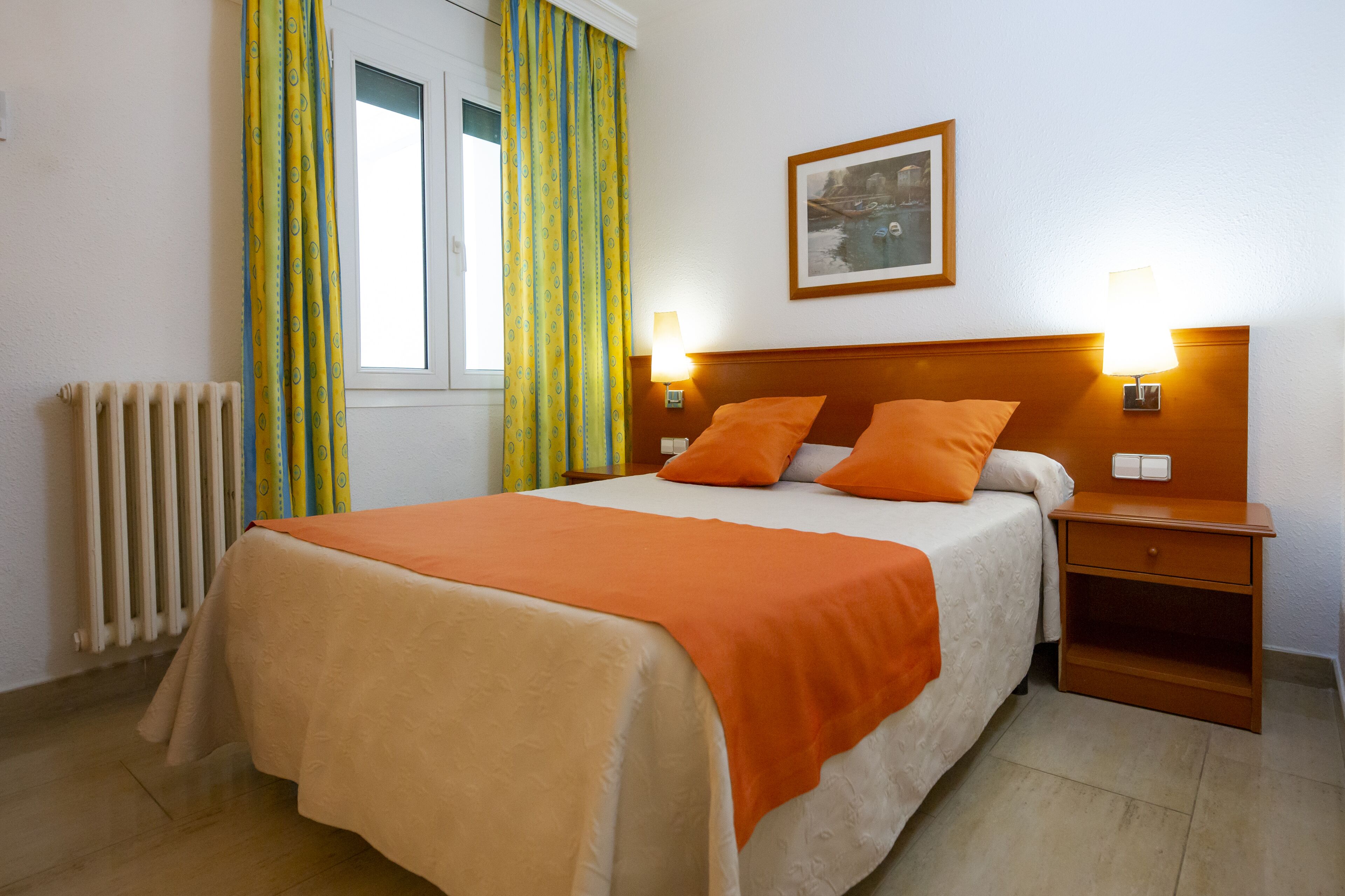 Una cama bien tendida con una colcha naranja, dos lámparas de noche, una mesita de noche con cajón y una ventana con cortinas.