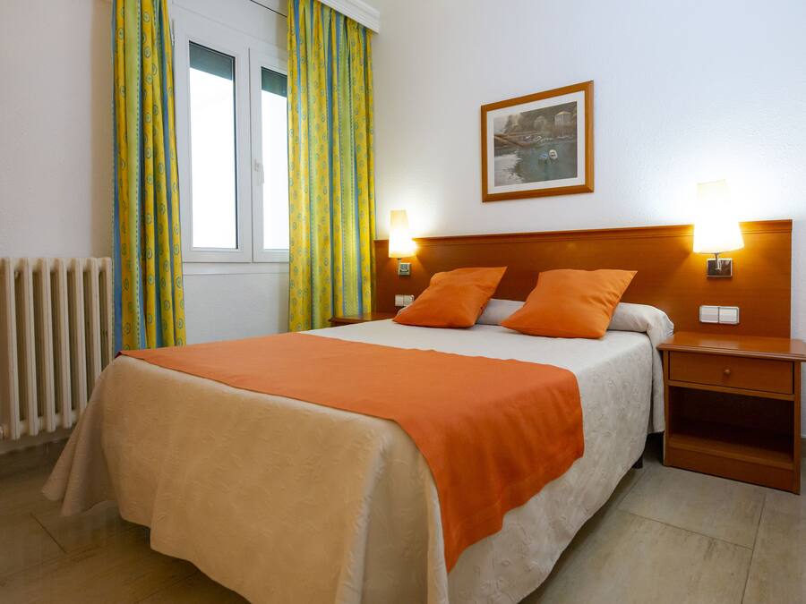 Una cama bien tendida con una colcha naranja, dos lámparas de noche, una mesita de noche con cajón y una ventana con cortinas.