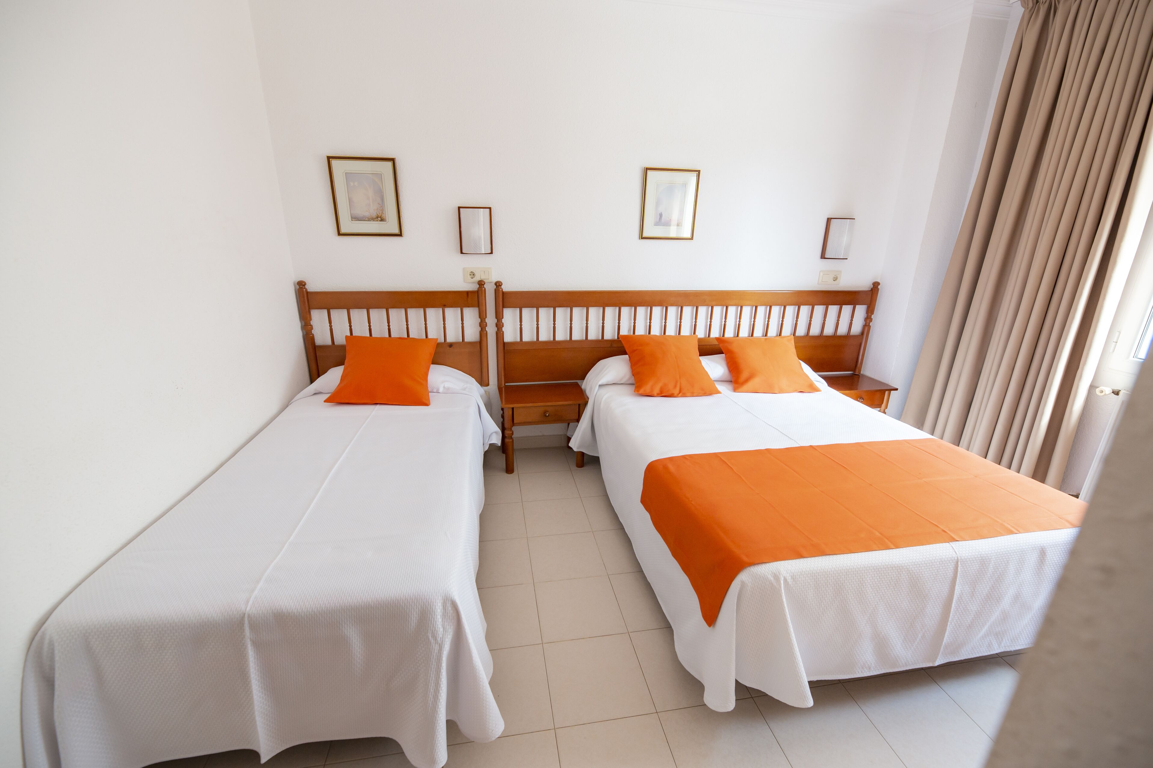 Habitación de hotel con dos camas individuales, cada una con una colcha y almohada naranja, ropa de cama blanca y un cabecero de madera. Se observan cuadros enmarcados en la pared y una ventana con cortinas.