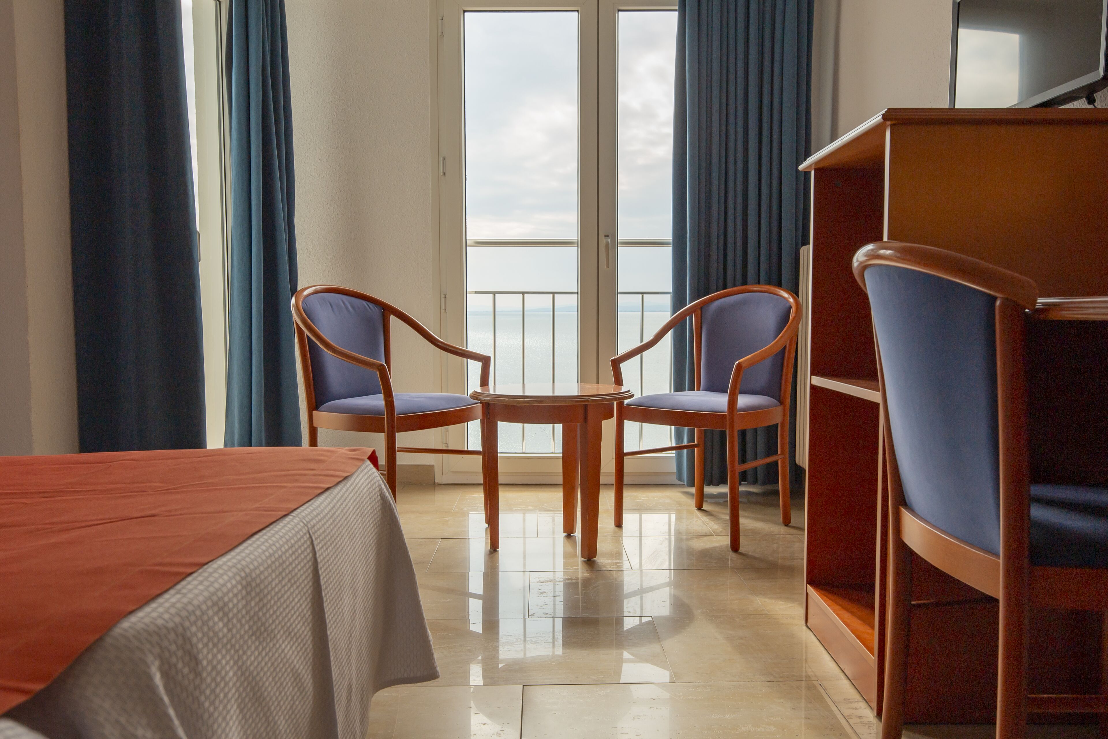 A hotel room with a bed, two chairs, a small table, and a bookshelf.