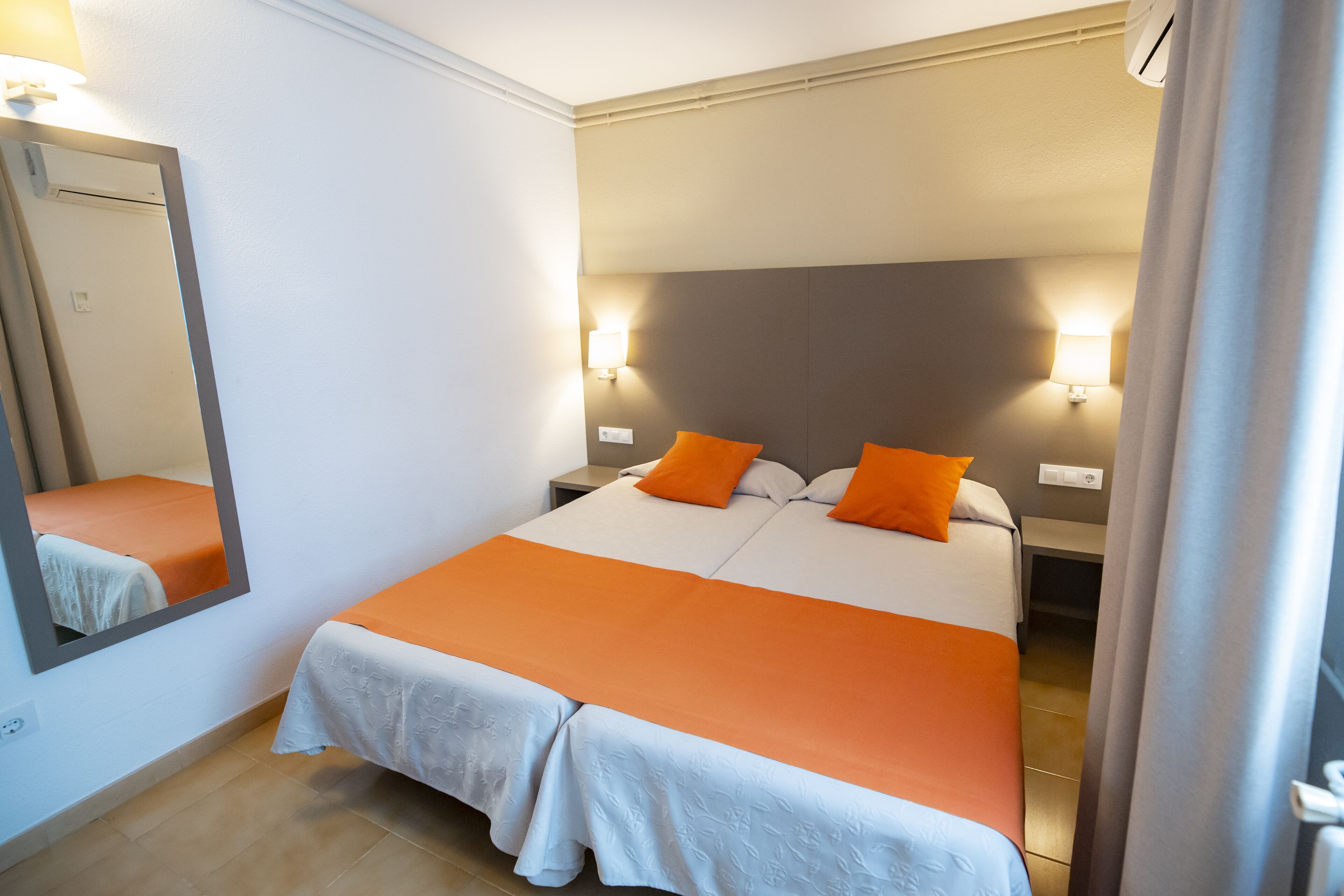 Una cama doble con ropa de cama naranja, una mesita de noche, una lámpara de pared y un espejo.