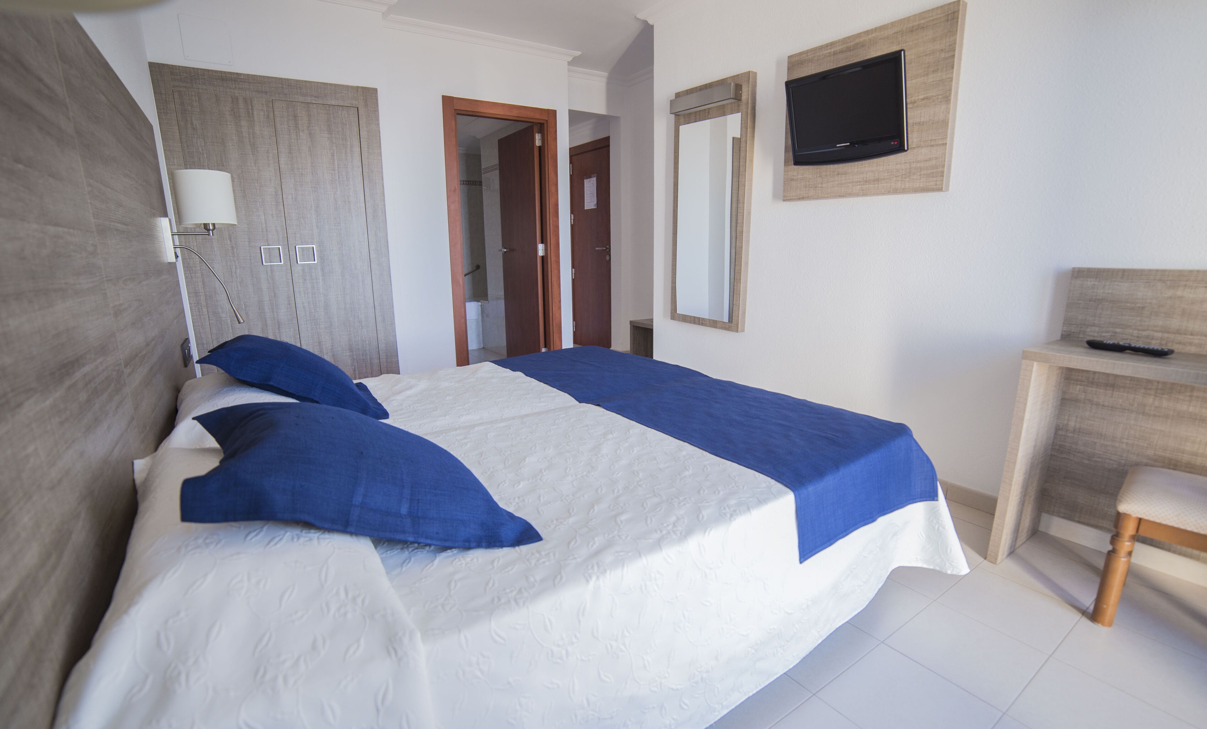 A neatly made bed with blue and white bedding, a wooden headboard, a bedside lamp, a TV mounted on the wall, and a wooden desk with a chair.
