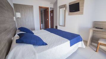 Una cama bien hecha con ropa de cama azul y blanca, cabecera de madera, lámpara de noche, televisor montado en la pared y un escritorio de madera con una silla.