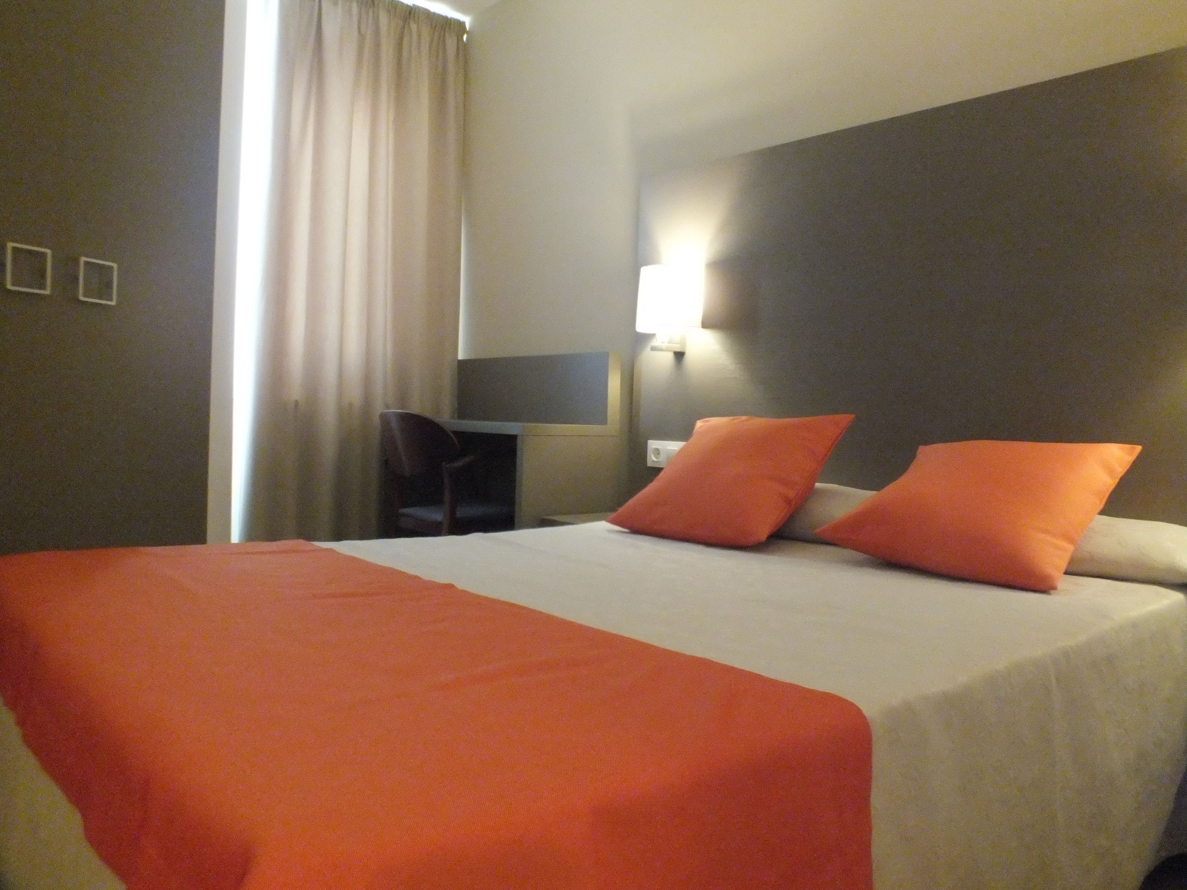 A hotel room with a bed, two orange pillows, a desk, and a chair.