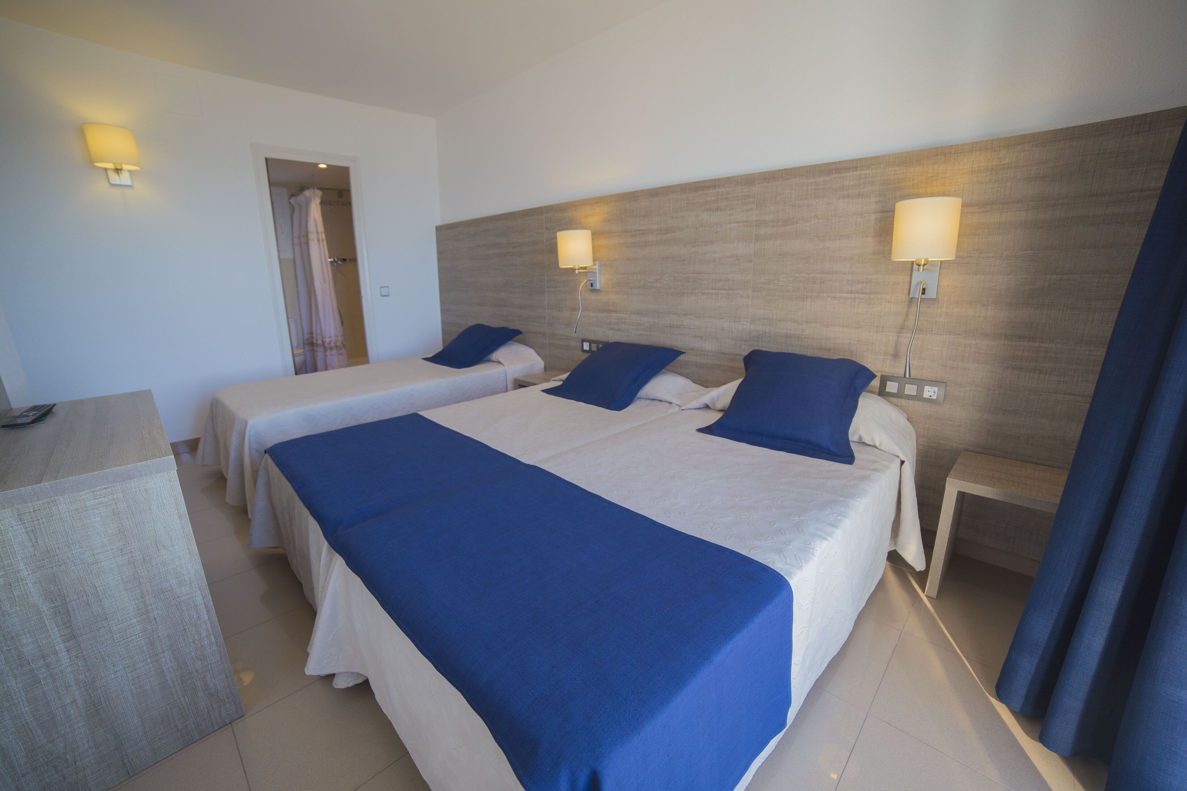 Habitación de hotel con dos camas, cada una con cubrecama y almohadas azules, cabeceros de madera, lámparas de noche y un escritorio con silla.