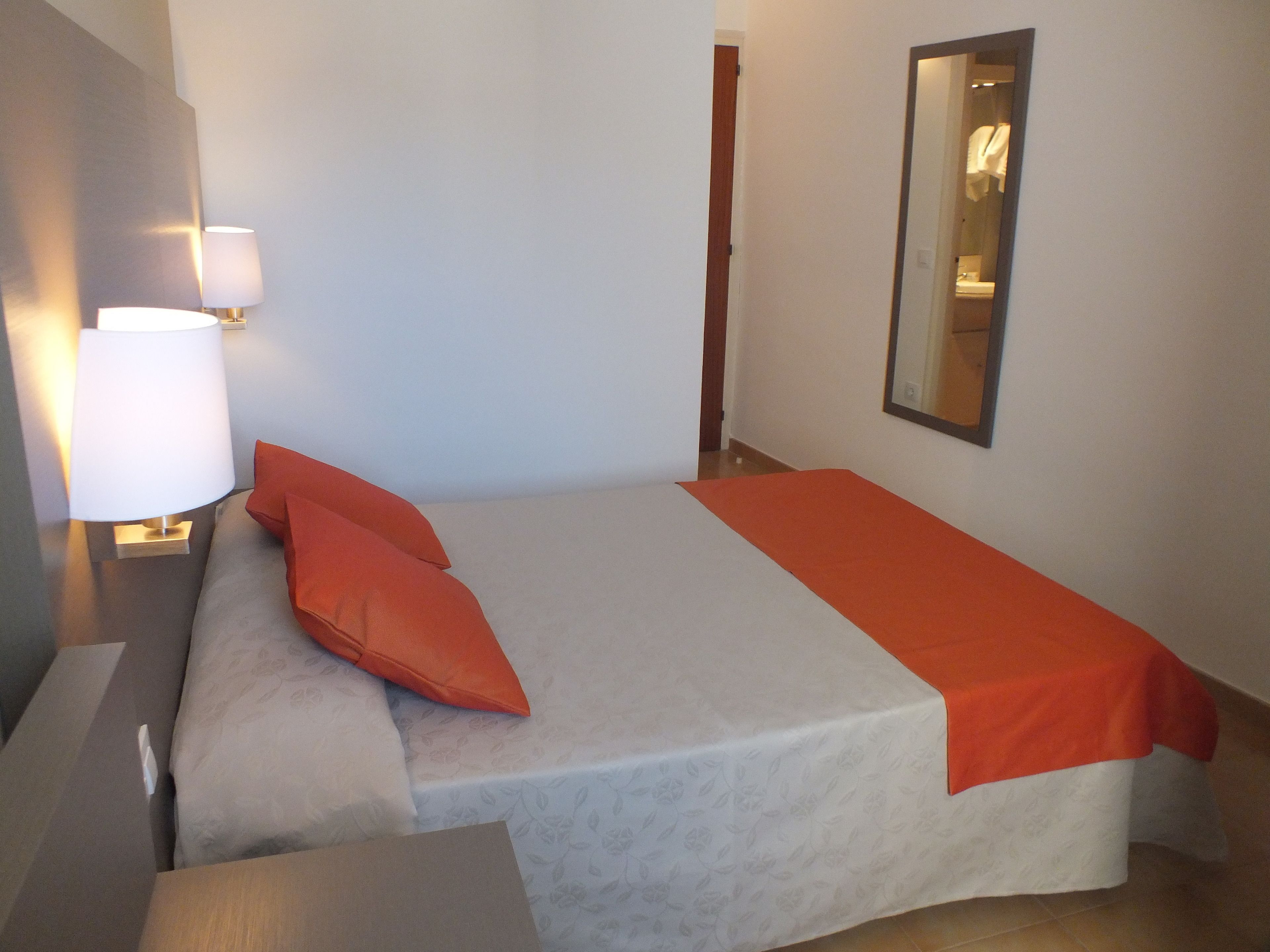 A neatly made bed with a white and orange bedspread, a single orange pillow, and a bedside lamp.