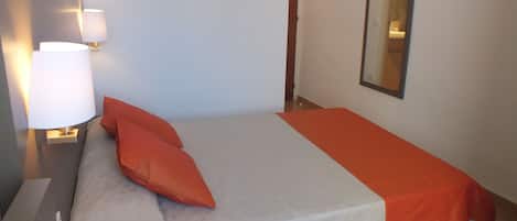 Una cama bien tendida con una colcha blanca y naranja, una almohada naranja y una lámpara de noche.