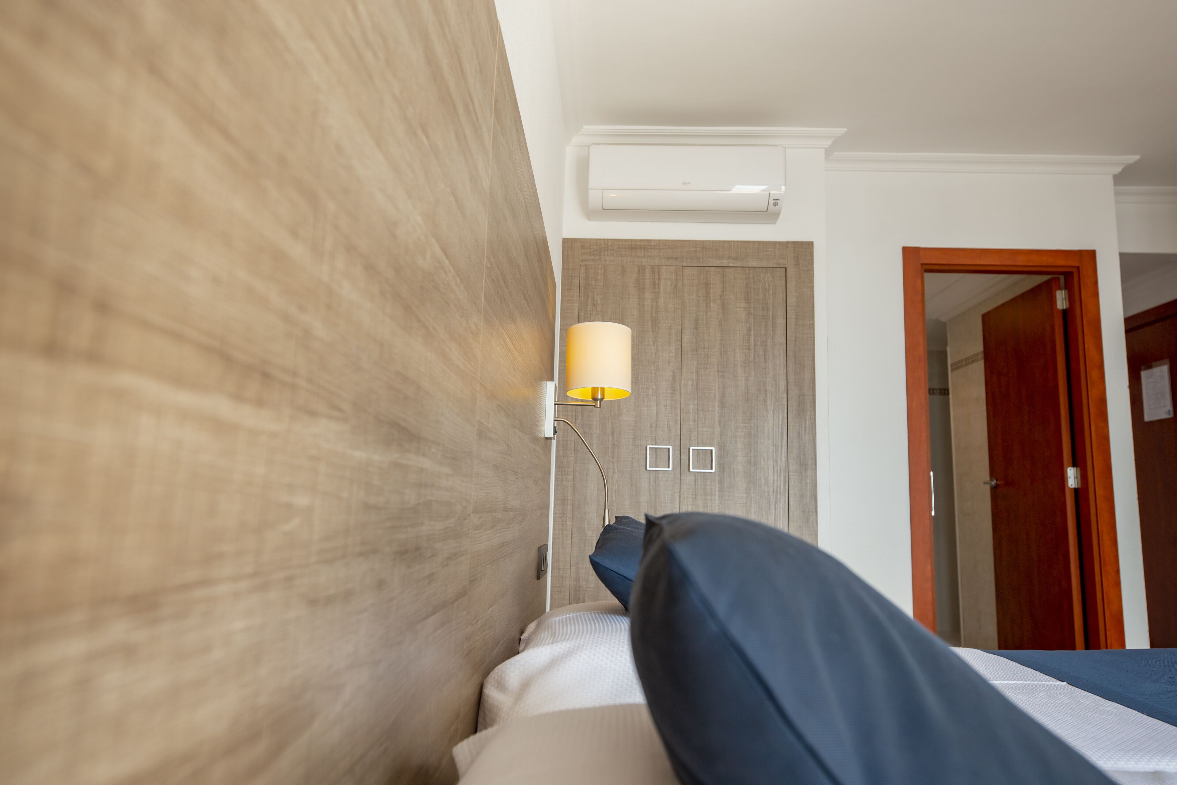 A hotel room with a wooden headboard, a bedside lamp, a bed with a blue cover, a wooden door, and an air conditioning unit on the wall.