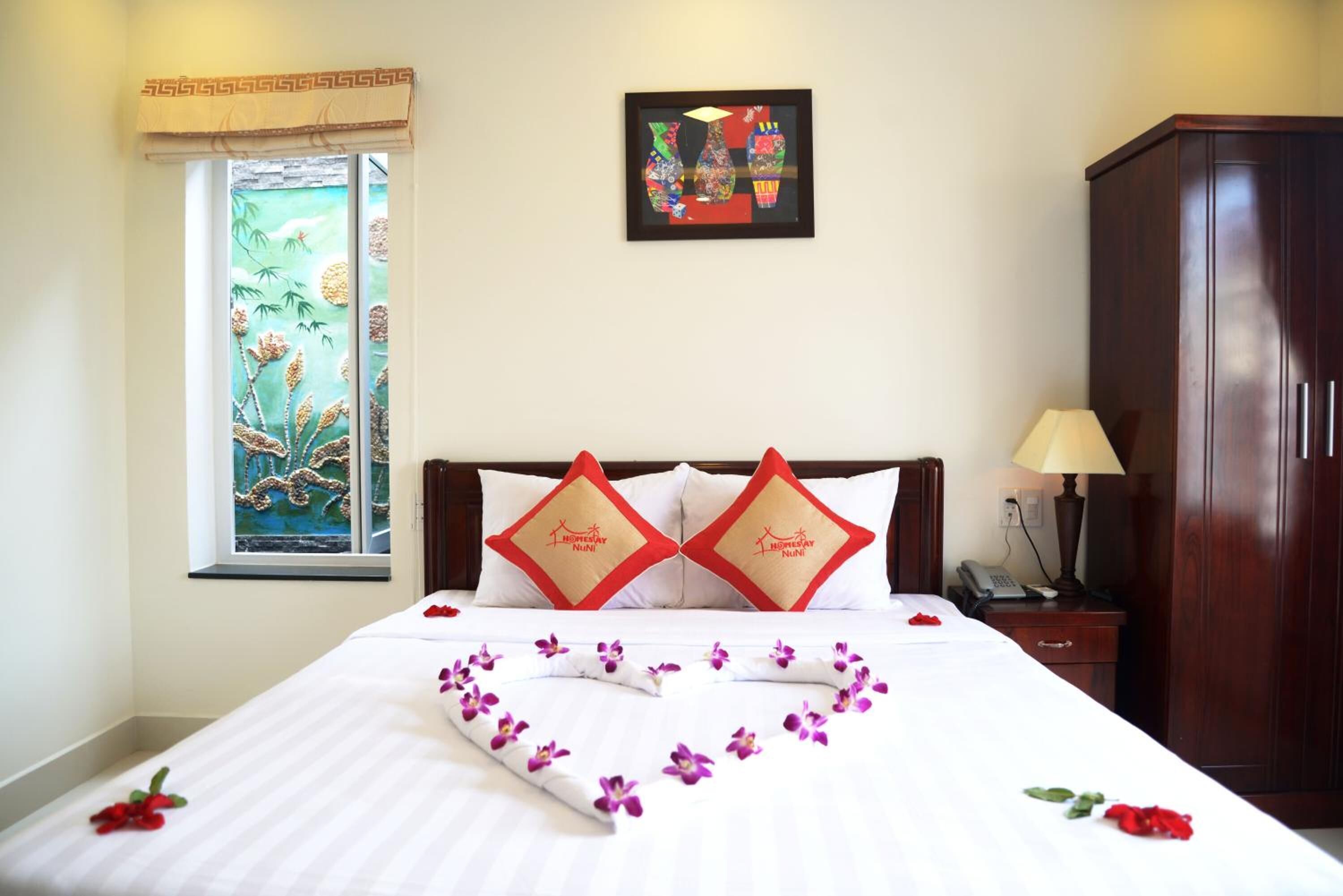 A hotel room with a bed, a window with a view of bamboo, a painting on the wall, and a wooden wardrobe.