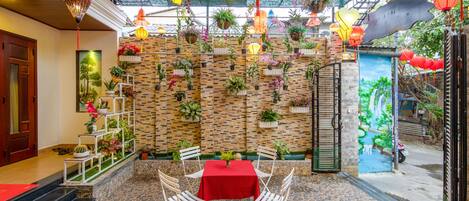 Un patio con una mesa y sillas rojas, rodeado de plantas en macetas y faroles colgantes.