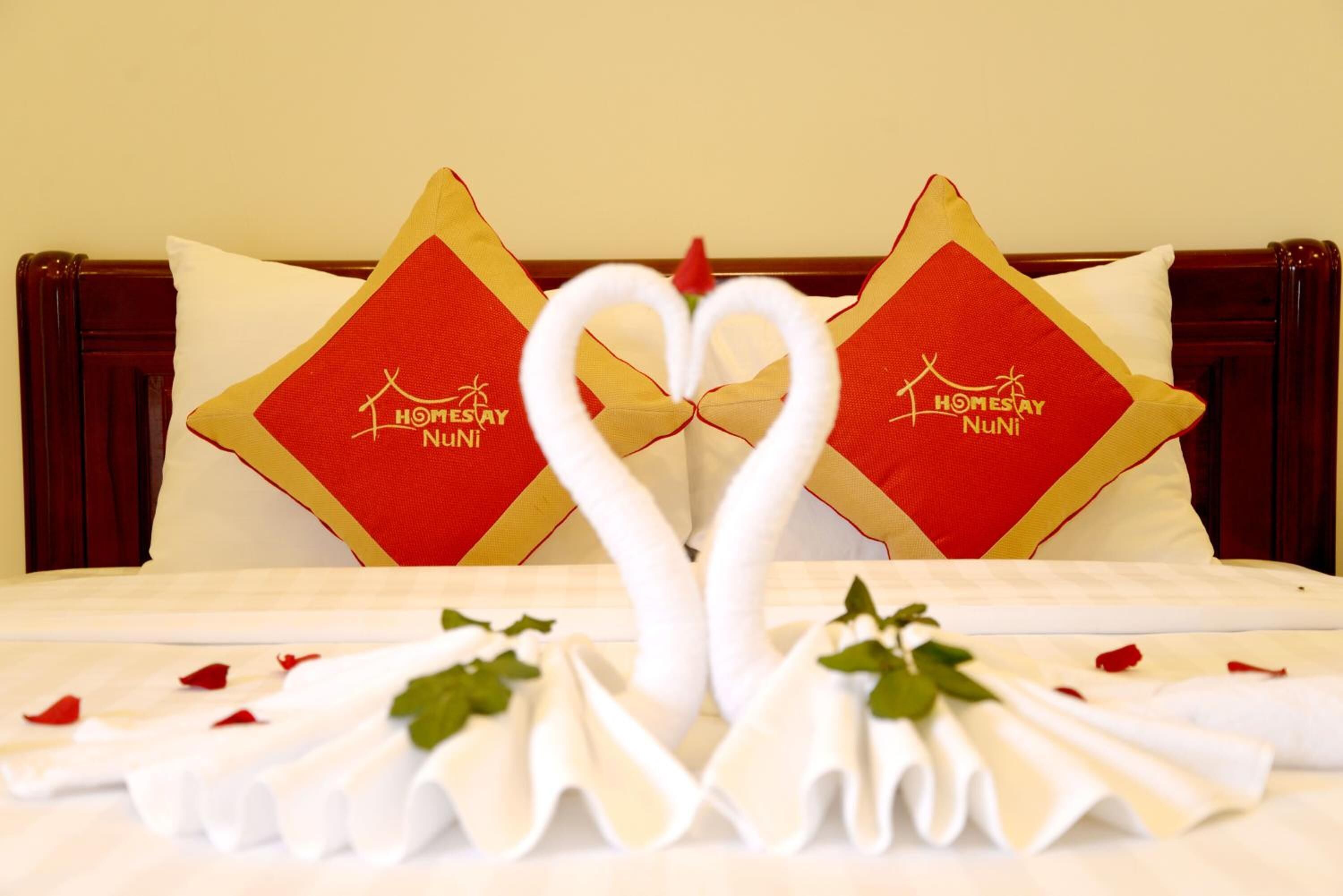 A hotel room with a bed, two pillows with a red and gold design, and a heart-shaped arrangement of white towels and rose petals.