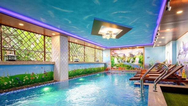 An indoor swimming pool with a mural of a garden on the walls, a ceiling with a water effect, and a poolside area with lounge chairs and a sunshade. - NuNi Hoi An Homestay (Da Nang)