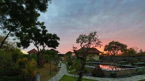 A garden with a pathway leading to a building with a red roof, surrounded by trees and a small pond. - Segara Tegeh - Hostel (Tulamben)