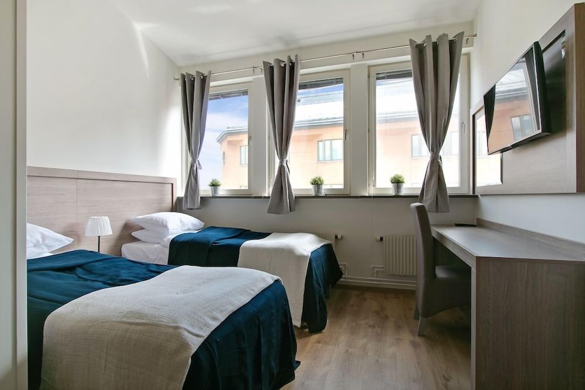 A hotel room with two beds, a desk, a TV, and a window with curtains.
