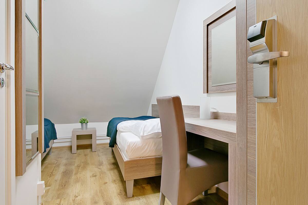 Ein Schlafzimmer mit Bett, Schreibtisch, Stuhl, einem kleinen Tisch mit Pflanze und Spiegel an der Wand.